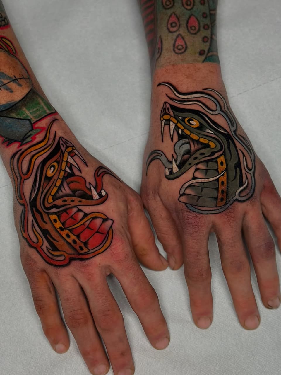 Two hands displaying matching dragon head tattoos in red, orange, yellow, and black with detailed scales and flowing hair elements on the knuckles and back of hands.