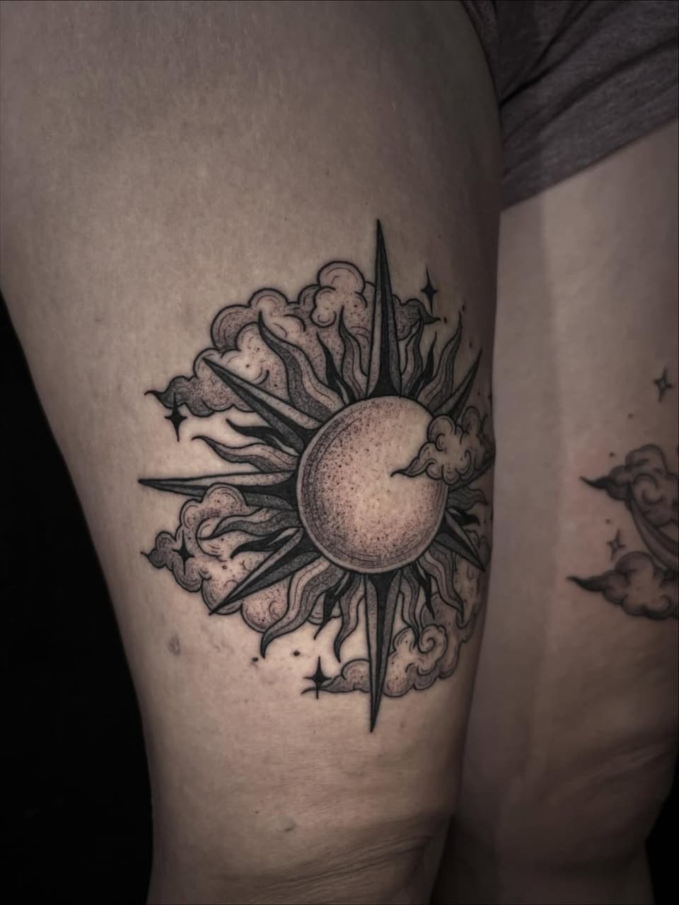 Decorative sun tattoo with radiating spikes and cloud-like petals on the thigh, rendered in black ink with dotted shading details.