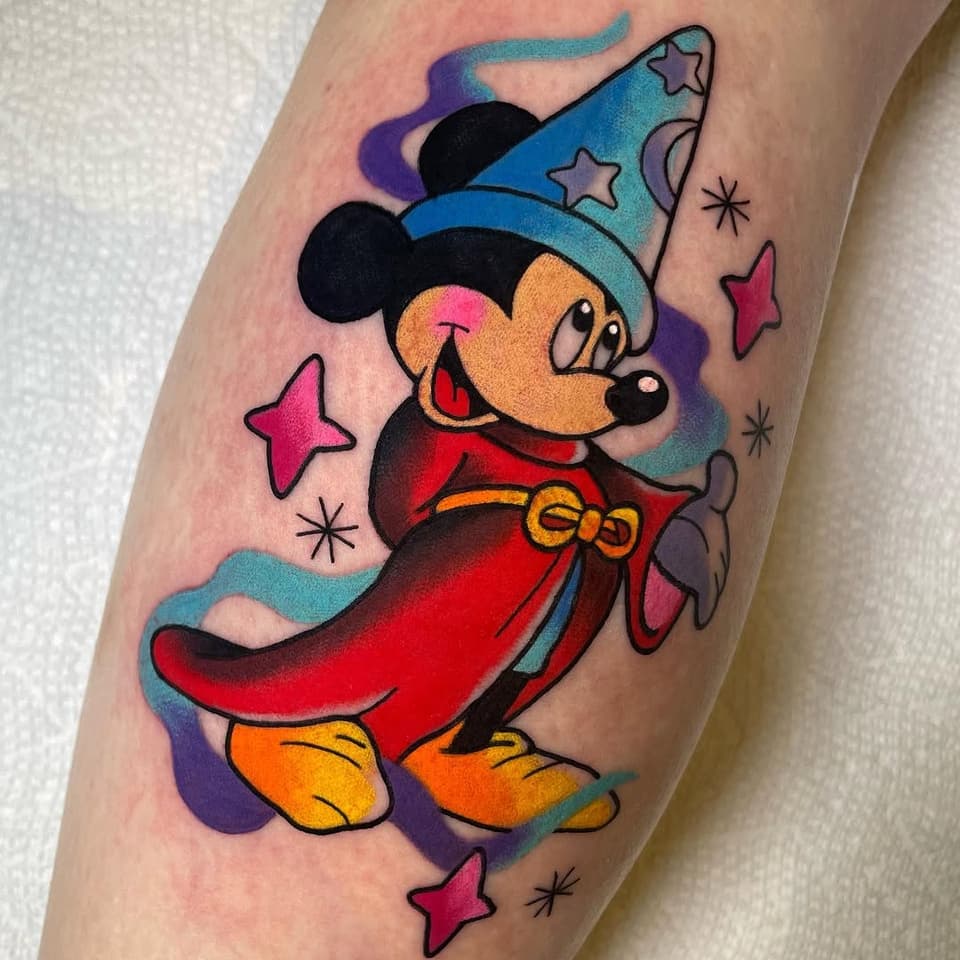 Colorful Mickey Mouse tattoo wearing a blue wizard hat and red cape, surrounded by pink stars and sparkles on the upper arm.