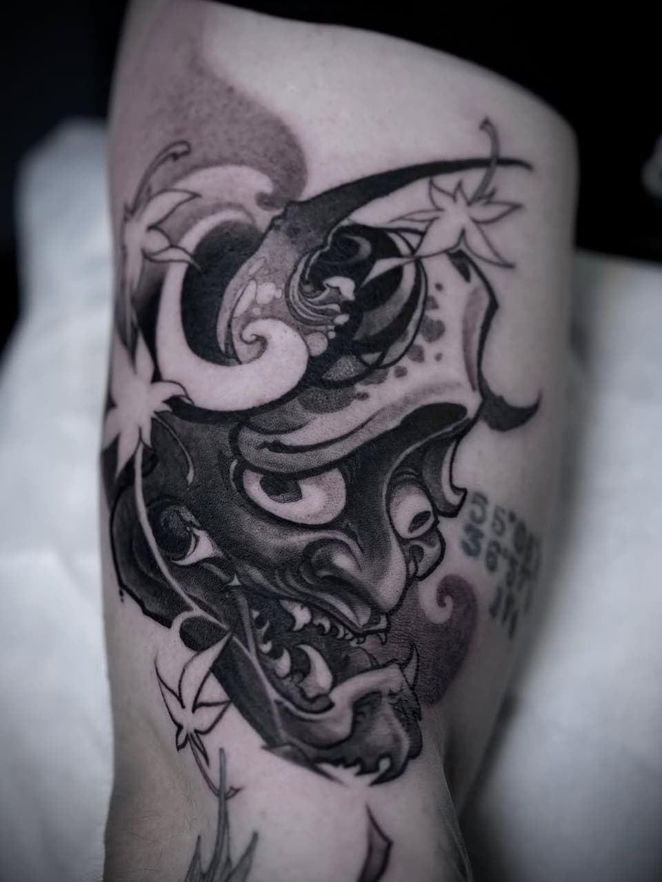 A detailed black and grey tattoo on the thigh features a horned demon face with ornate swirls, flowers, and decorative elements covering the upper leg.