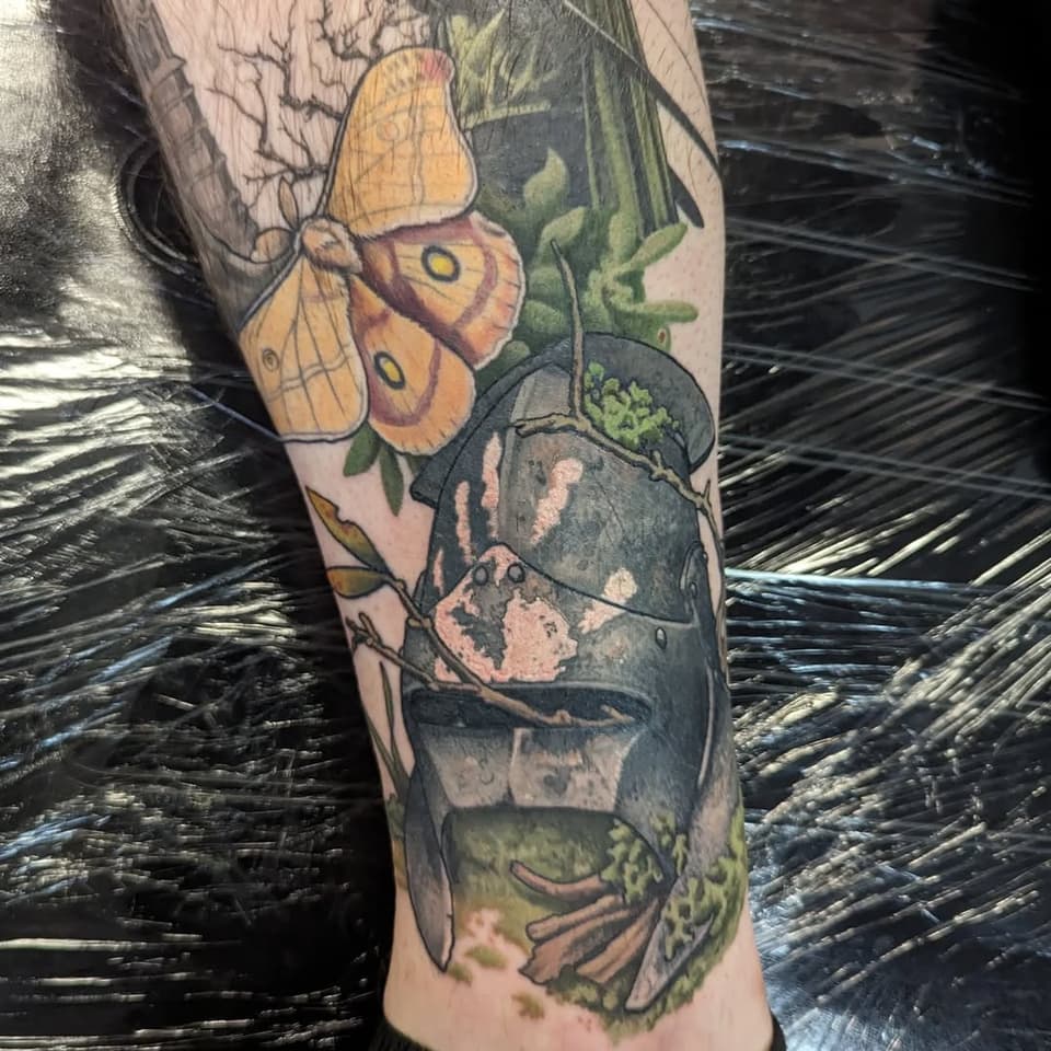 Colorful tattoo on leg featuring yellow and orange butterfly, dark moth, green leaves, and botanical elements with detailed shading.